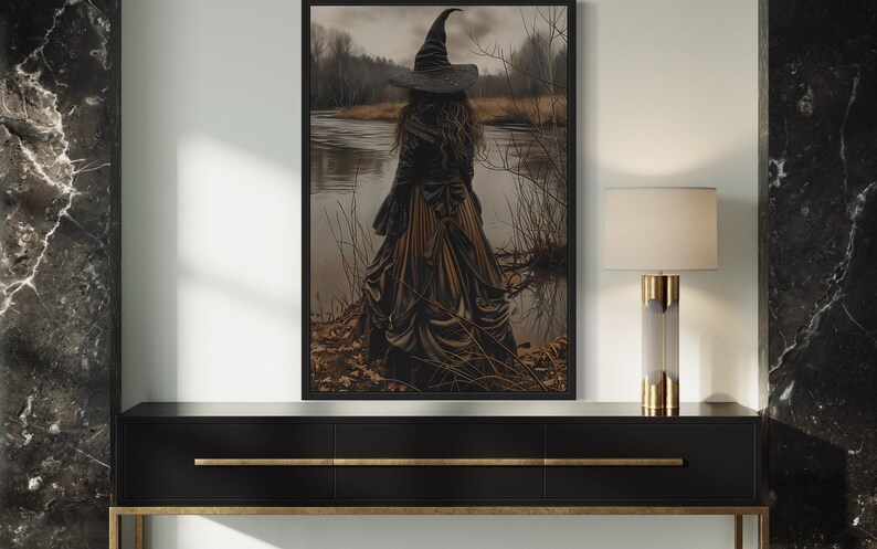 a large framed artwork depicting a woman in a long dress standing by a lake, with a witch's hat on her head. The artwork is hanging on a wall, and there is a black console table below it.