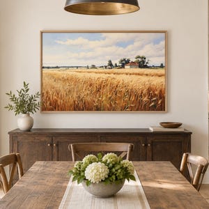 May include: A rectangular landscape painting in a light wood frame depicting a golden wheat field under a blue sky with white clouds, featuring a small farmhouse in the distance, displayed on a wall above a dark wooden sideboard in a dining room.