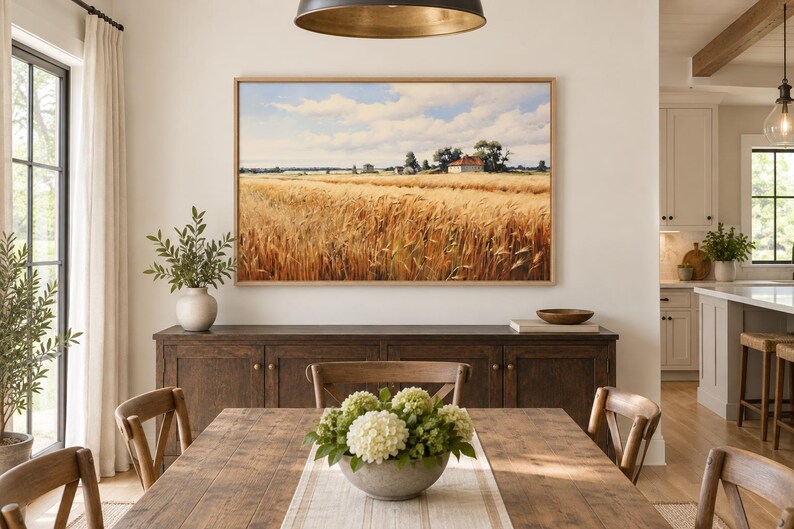 May include: A rectangular landscape painting in a light wood frame depicting a golden wheat field under a blue sky with white clouds, featuring a small farmhouse in the distance, displayed on a wall above a dark wooden sideboard in a dining room.