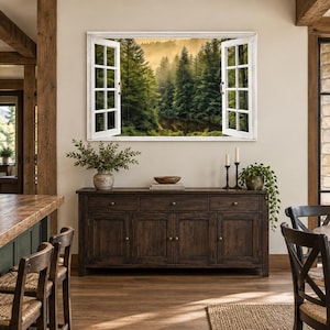 May include: A rectangular wall art piece featuring a white window frame with open shutters, displaying a scenic view of a dense, misty green forest. The artwork is mounted on a light beige wall above a dark brown wooden sideboard cabinet.