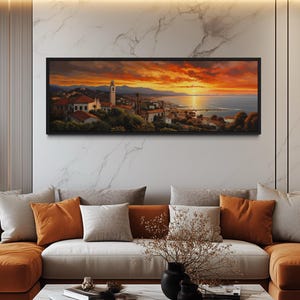 a large, framed painting of a picturesque coastal town at sunset, hanging on a wall above a couch with orange and gray pillows.