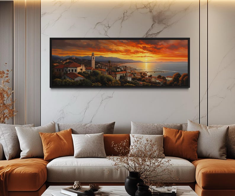 a large, framed painting of a picturesque coastal town at sunset, hanging on a wall above a couch with orange and gray pillows.