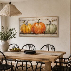 May include: A rectangular wall art piece featuring a painting of three pumpkins in yellow, orange, and muted green, set against a rustic white wood-plank background, enclosed in a light-toned wooden frame, displayed horizontally on a wall.