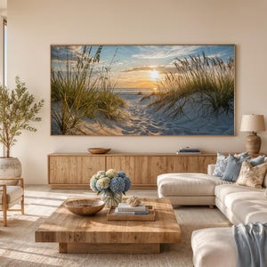 May include: A large rectangular wall art print featuring a sunset over a sandy beach with tall dune grasses, displayed in a light wood frame above a long wooden media console in a living room with a beige sectional sofa and a wooden coffee table.