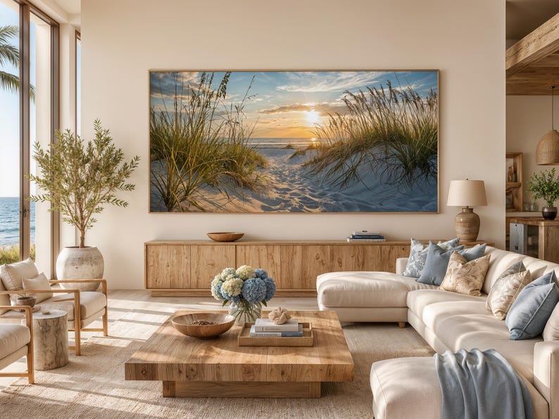May include: A large rectangular wall art print featuring a sunset over a sandy beach with tall dune grasses, displayed in a light wood frame above a long wooden media console in a living room with a beige sectional sofa and a wooden coffee table.