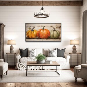 a cozy living room with a large painting of three pumpkins hanging on the wall. The room is furnished with a white sofa, two armchairs, and a coffee table. The room is decorated with a rustic wooden floor, a chandelier, and a window with curtains.