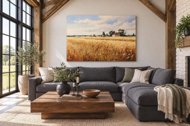 May include: A large rectangular landscape painting depicting a golden wheat field under a blue sky with white clouds, mounted on a white wall above a dark gray sectional sofa, a wooden coffee table, and a woven rug in a bright living room.