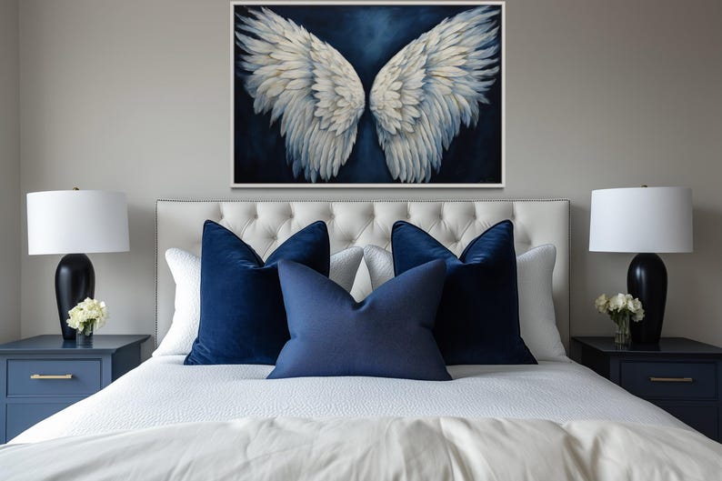 a bedroom with a large bed, white bedding, and blue accent pillows. Above the bed is a framed artwork of white and blue angel wings. On either side of the bed are nightstands with black lamps, and there are white flowers on the nightstands.