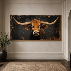 May include: A large rectangular wall art piece featuring a detailed portrait of a brown longhorn cow with prominent horns. The animal is set against a dark, textured background with hints of gold and bronze tones, framed in a thin gold frame.