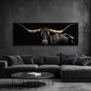 May include: A panoramic wall art print featuring a longhorn steer with dark fur and prominent, wide-spanning horns against a solid black background, framed in a thin black border and mounted horizontally on a dark grey wall above a sofa.