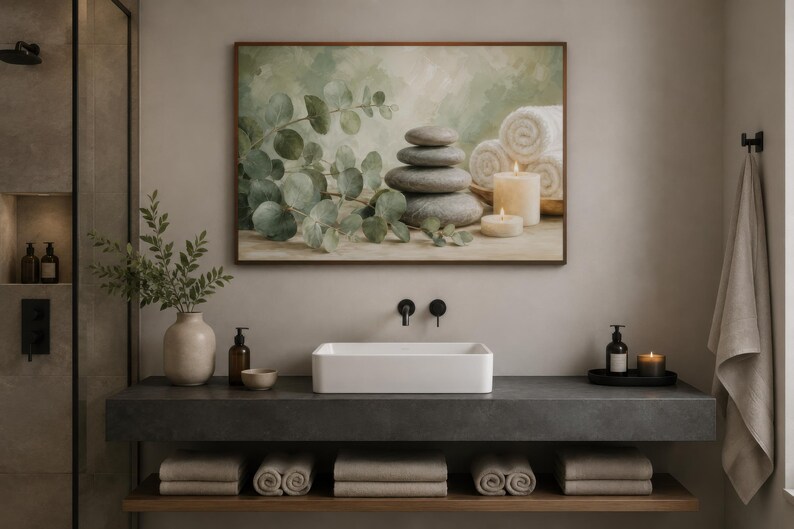 May include: A rectangular wall art print featuring a stack of smooth grey stones, green eucalyptus branches, white rolled towels, and two lit pillar candles. The artwork is displayed in a thin brown frame above a white rectangular bathroom sink.