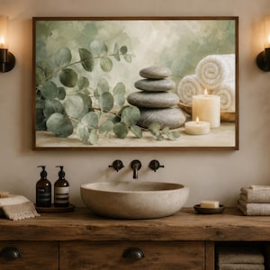 May include: A rectangular wall art print featuring a stack of smooth grey stones, green eucalyptus branches, rolled white towels, and two cream-coloured pillar candles. The artwork is displayed in a wooden frame above a bathroom vanity with a stone sink.