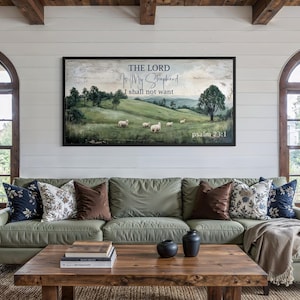 Könnte beinhalten: Ein Wohnzimmer mit einem salbeigrünen Sofa und dekorativen Kissen. Ein Couchtisch aus Holz zeigt Bücher und Ornamente. Über dem Sofa hängt ein gerahmtes Landschaftskunstwerk mit dem Text "The Lord is My Shepherd, I shall not want."