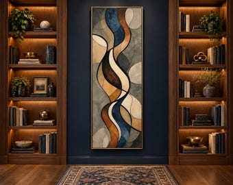 May include: A tall, rectangular framed wall art piece featuring an abstract design of flowing, organic shapes in shades of navy blue, warm brown, cream, and muted grey, displayed on a dark blue wall between two illuminated wooden bookshelves.