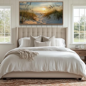 a serene bedroom with a large bed, a nightstand, and a painting of a beach scene above it. The room has large windows that provide a view of the outdoors, and the overall decor has a neutral color palette with a touch of natural elements.
