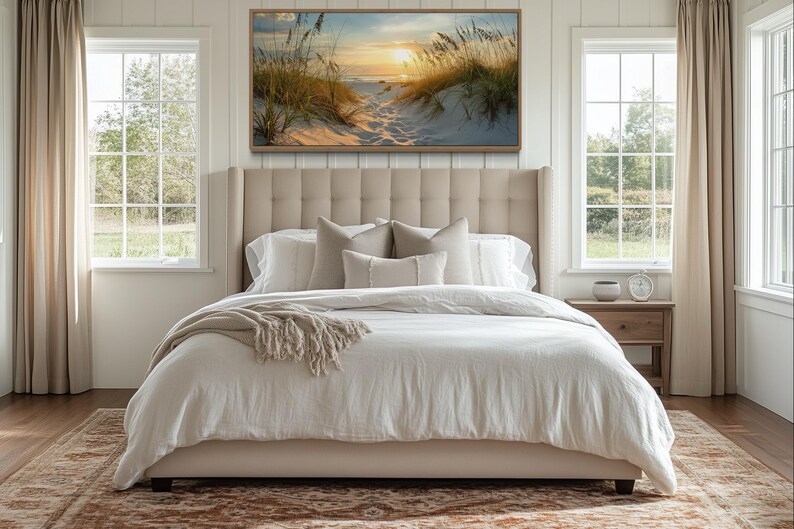 a serene bedroom with a large bed, a nightstand, and a painting of a beach scene above it. The room has large windows that provide a view of the outdoors, and the overall decor has a neutral color palette with a touch of natural elements.