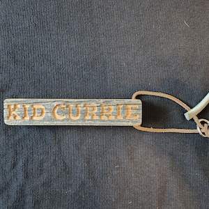 May include: A wooden keychain with the text "KID CURRIE" carved into it. The keychain is attached to a brown leather cord and a metal hook.