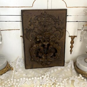 Peut inclure: Plaque rectangulaire en métal bronze antique avec un motif en relief orné. Le relief présente un blason central avec des motifs tourbillonnants et floraux. Le tout est posé sur un fond blanc avec des éléments décoratifs.