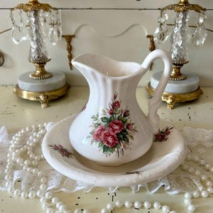 May include: White ceramic jug and basin set with a pink and green rose floral design. The jug has a curved handle and sits on a matching basin. Two crystal candle holders are in the background.