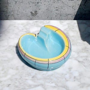 May include: A ceramic ashtray with a light blue interior and a curved rim featuring yellow, pink, and blue segments separated by thin black grid lines. The bowl has a central indentation for resting items, with a smooth, glossy finish throughout.