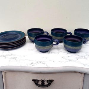 May include: A set of ceramic tableware featuring a stack of plates and five matching teacups. The plates and cups are a deep blue with a gradient of teal and purple. The set is arranged on a white marble-patterned surface.