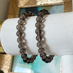 May include: Two smoky quartz beaded bracelets. Each bracelet features round, translucent beads in shades of gray and brown. The bracelets are displayed on a white surface, with a gold-framed object in the background.