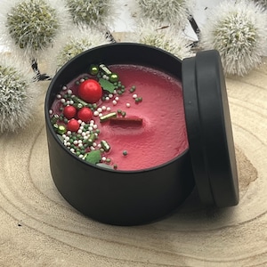 Könnte beinhalten: Eine rote Kerze in einer schwarzen Dose mit Dekorationen. Die Kerze ist mit roten und grünen Streuseln und einem Holzdocht verziert. Die Dose ist geöffnet und zeigt die Kerze. Im Hintergrund sind Pusteblumen zu sehen.
