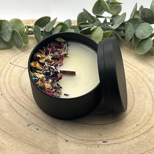 Könnte beinhalten: Eine schwarze Kerzendose mit teilweise geöffnetem Deckel. Die Kerze ist mit weißem Wachs gefüllt und mit getrockneten Blumen und einem Holzdocht verziert. Die Kerze steht auf einer Holzoberfläche.