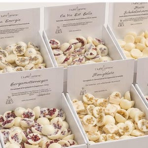 Pode incluir: Dez caixas de cartão branco dispostas em duas filas, cada uma cheia de pequenos doces redondos cobertos com chocolate branco. Cada caixa apresenta um rótulo impresso com texto e um pequeno logótipo. Um ramo de eucalipto verde é visível à esquerda.