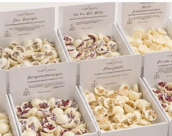 Op de afbeelding: Tien witte kartonnen doosjes in twee rijen, gevuld met diverse kleine, ronde, met witte chocolade omhulde lekkernijen. Elk doosje heeft een bedrukt etiket met tekst en een klein logo. Een groene eucalyptus tak is links zichtbaar.