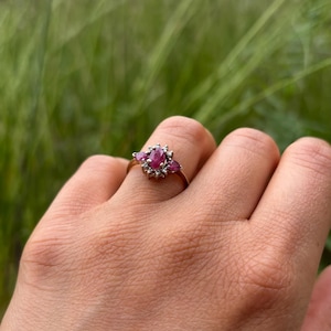 Könnte beinhalten: Ein goldener Ring mit drei ovalen, tiefrosa Edelsteinen, umgeben von kleineren, klaren Steinen. Der Ring ist auf einem zarten Goldband gefasst und zeigt ein klassisches Design. Der Ring wird an einer Hand getragen.
