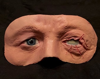 May include: A flesh-toned prosthetic mask featuring a human eye and surrounding skin. One eye is blue and open, the other is closed with visible lacerations and stitches. A realistic horror prop.
