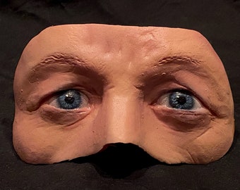 May include: A close-up of a realistic-looking face mask with blue eyes and a sculpted nose. The mask is made of a flesh-toned material and appears to be a prop for a play or film.