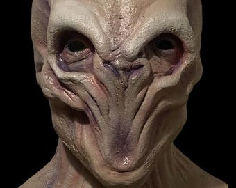 May include: A latex alien mask with a textured, pale skin tone. The mask features large, dark eyes, a prominent brow, and a long, thin nose. The mask's design includes detailed facial features and a neck piece.