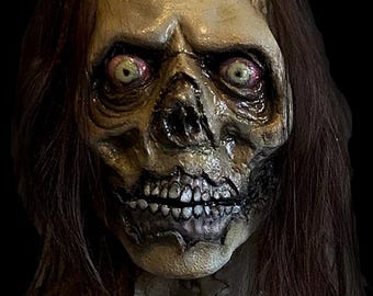 May include: A highly detailed zombie mask with visible teeth, sunken eyes, and decaying skin. The mask has long, dark brown hair and is set against a black backdrop, ideal for Halloween.