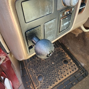 May include: A close-up of a black gear shift knob in a car interior. The knob is round and has a textured surface. The car's dashboard is visible in the background, with various buttons and controls.