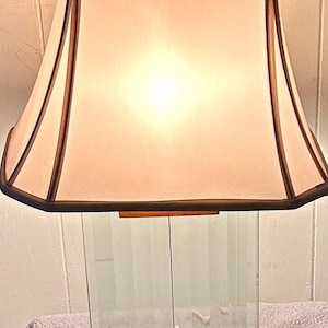May include: A rectangular table lamp with a clear glass base and a beige lampshade. The lampshade has a dark brown trim and a rectangular shape. The base has a wooden accent. The lamp is turned on, and the light is shining through the lampshade.