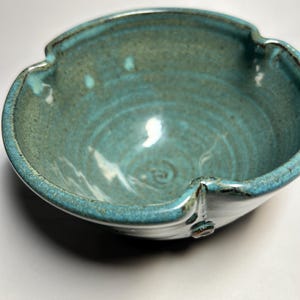 May include: A handmade ceramic bowl with a unique, scalloped rim. The bowl is glazed in shades of teal and turquoise, with darker accents around the rim. The interior shows spiral patterns. A decorative piece for the kitchen or dining room.