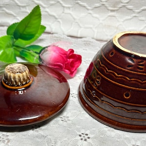 May include: A brown ceramic bowl and lid with a decorative design. The lid has a textured handle. A pink rose and green leaves are in the background. The bowl is approximately 6 inches in diameter.