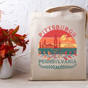 May include: A beige tote bag with a retro-style design featuring a cityscape of Pittsburgh, Pennsylvania. The design includes a sunset, the words "Pittsburgh" and "Pennsylvania", and a football helmet with the word "Steelers".