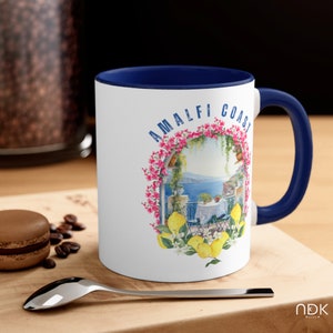 May include: White ceramic mug with a blue rim and handle. The mug features a colorful illustration of a lemon tree with lemons and flowers, a view of the ocean, and the text "Amalfi Coast".