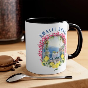 May include: White ceramic mug with a black rim and handle. The mug features a colorful illustration of a floral archway with a view of the Amalfi Coast and the text "Amalfi Coast".