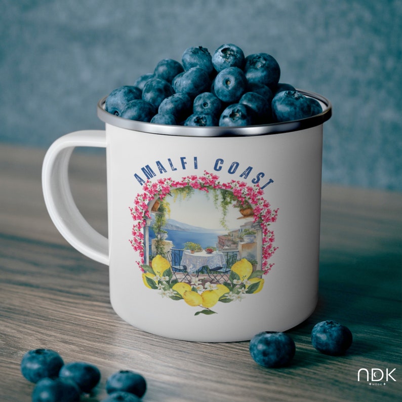 May include: White enamel mug with a blue rim and handle. The mug has a colorful illustration of a coastal scene with the text "Amalfi Coast" printed on it. The mug is filled with blueberries.