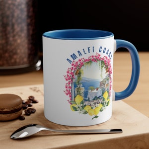 May include: White ceramic mug with a blue rim and handle. The mug features a colorful illustration of a lemon tree with lemons and flowers, a view of the ocean, and the text "Amalfi Coast".