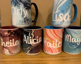 May include: Six personalized coffee mugs with marbleized designs. The mugs are in various colors, including blue, pink, and brown. The names "Orly", "Lisa", "Sheila", "Alicia", "Hailey", and "May" are written in white on the mugs.