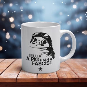 Peut inclure: Un mug en céramique blanche orné d'une illustration graphique noire représentant un cochon portant des lunettes d'aviateur et une écharpe. Sous l'illustration, le texte "BETTER A PIG THAN A FASCIST" est écrit en gras, suivi d'une étoile.