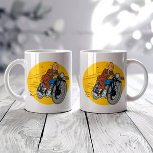 Puede incluir: Dos tazas de cer&aacute;mica blanca con una ilustraci&oacute;n de dibujos animados de un hombre en motocicleta. La ilustraci&oacute;n presenta un fondo amarillo con el hombre con traje marr&oacute;n y camisa azul. La moto es negra y gris.