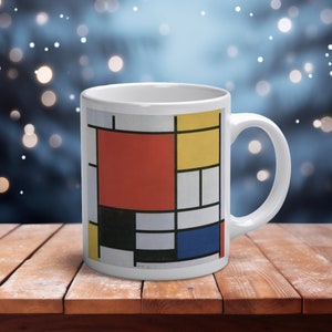 May include: A white ceramic mug featuring a geometric grid pattern composed of thick black lines and rectangular blocks of primary colors including red, yellow, and blue, set against a white background with a curved handle on the right side.