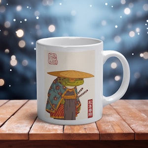 Peut inclure: Un mug en céramique blanche orné d'une illustration graphique représentant une grenouille verte portant un chapeau de paille traditionnel et un kimono bleu à motifs floraux, avec deux épées à la ceinture, du texte japonais et un sceau rouge.