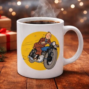 Puede incluir: Taza de caf&eacute; de cer&aacute;mica blanca con una ilustraci&oacute;n de dibujos animados de un hombre en una motocicleta sobre un fondo amarillo. La taza contiene caf&eacute; oscuro, del que sale vapor. La taza est&aacute; sobre una superficie de madera.